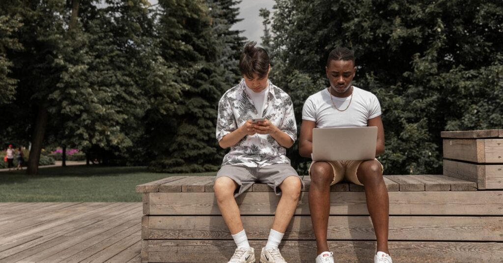 Zwei junge Männer nutzen im Park ein Smartphone und einen Laptop
